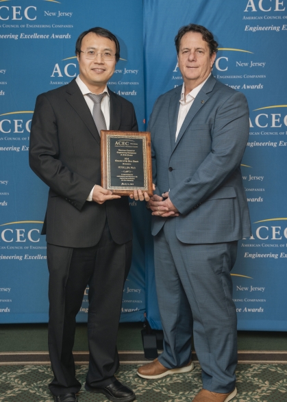 Two gentleman pose holding an award plaque between them. They are wearing suits and standing in front of a blue backdrop.